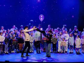 A large group of children and adults stand on stage, some watching two people in the center