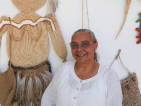 Photograph of Aunty Annette standing in front of her weaving artworks that are hung on a white wall.
