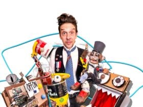 A man in a black vest and tie looking whimsically at the camera against a backdrop of toys