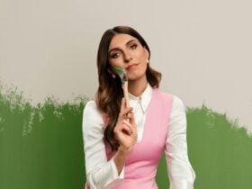 A woman with long brown hair in a white shirt and pink dress holds a paintbrush to her cheek