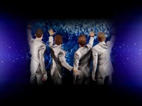 Four men in gray suits looking away from the camera into an audience, with a blue and black backing