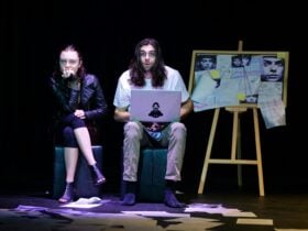 Two people seated, one holding a laptop with a pinboard covered in pictures