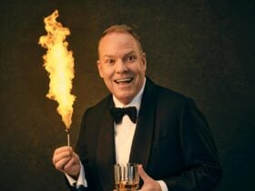 man in tuxedo holding a glass of whiskey and a lit match with a huge flame