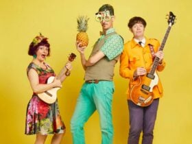 Three people stand against a yellow background. The left person holds a ukulele, the middle person w