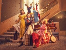 A group of five people in colorful 18th-century costumes stand on a staircase with instruments.