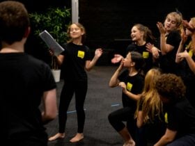 Children participating in a drama workshop stand barefoot on a studio floor, performing a scene toge
