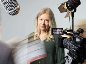 A woman with long blonde hair looks seriously at the camera, surrounded by filming equipment