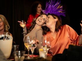 Two ladies sitting with heads together are singing happily holding glasses of wine
