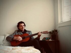 A man laying on a bed smiling at the camera, holding a guitar and wearing a suit