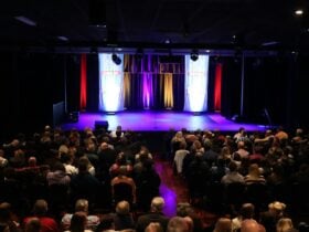 A crowd of theatre guests facing a lit stage with two Sydney Comedy Festival banners on it