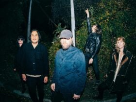 A group of five people stands in a dimly lit, wooded area. They wear casual, dark clothing.