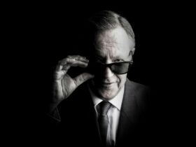 A man in a suit and tie adjusts his sunglasses while looking forward.