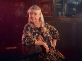 A woman with long gray hair sits at a round table in a dimly lit room, wearing a floral dress