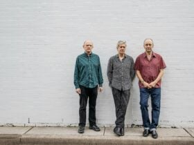 image of three men, standing infront of a grey wall. They are the three members of "The Necks"