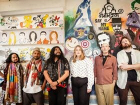 Six people stand side by side in front of a colorful mural featuring various faces and patterns.