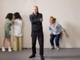 An older man standing with crossed arms with his three daughters in the background