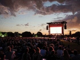 A large film screen is set up in wide open park space with festival goers looking at the screen