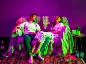 A man and woman in casual clothes laugh together on a couch under pink and green lighting.