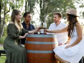 Four people sitting on a barrel outside, sharing a bottle of red wine.