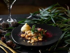 Elegant dish on a black plate with colorful garnishes, nuts, and puree. Surrounded by green leaves.