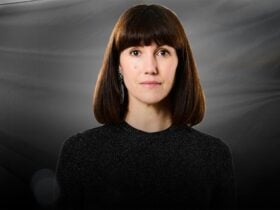 A woman with a long bob and bangs, staring blankly at the camera with a dark background