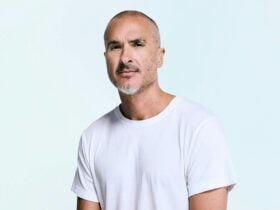 Man in a white t-shirt with a beard staring directly at the camera infront of light blue backdrop.