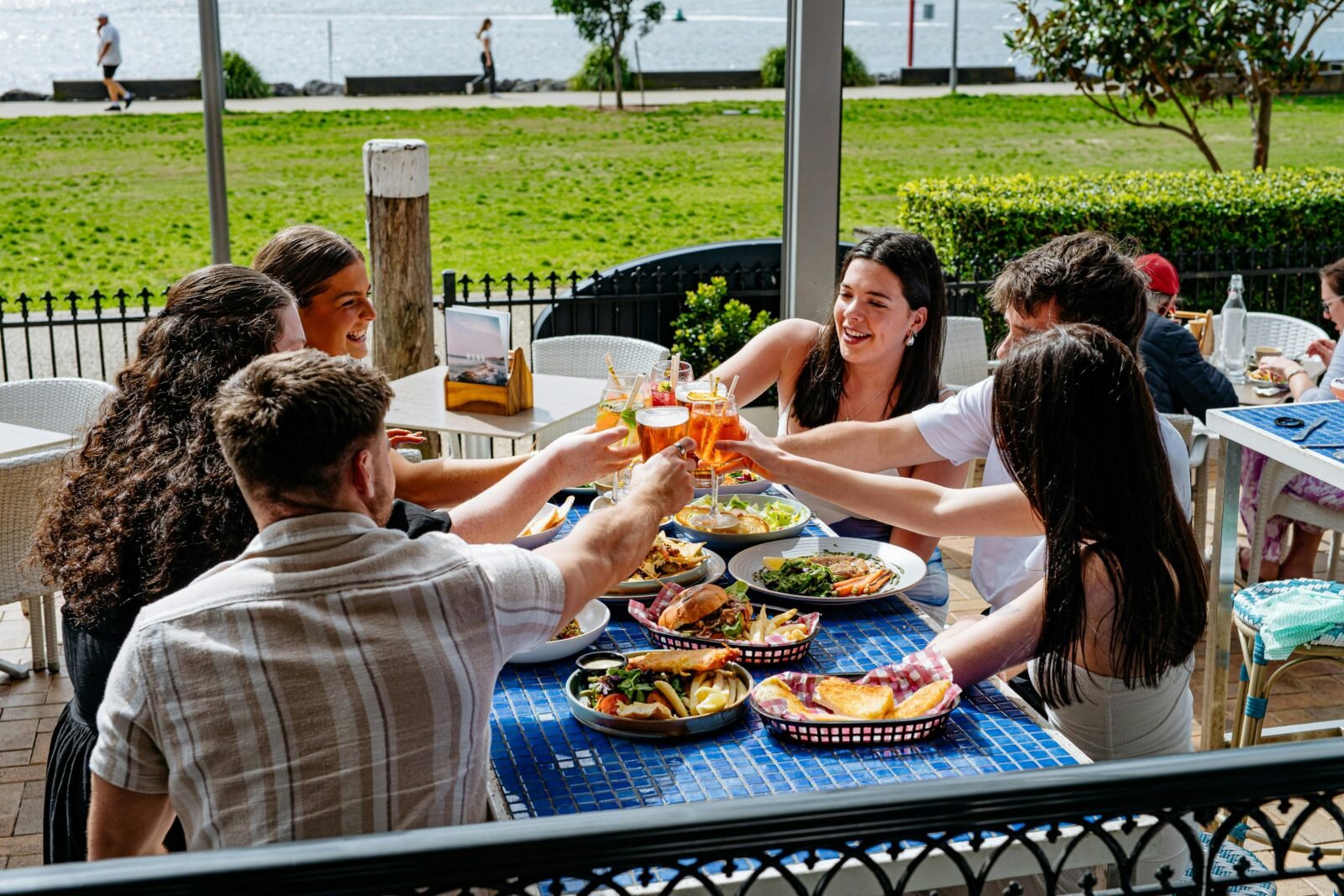 Friends clinking glasses over lunch at an outdoor table overlooking the water.