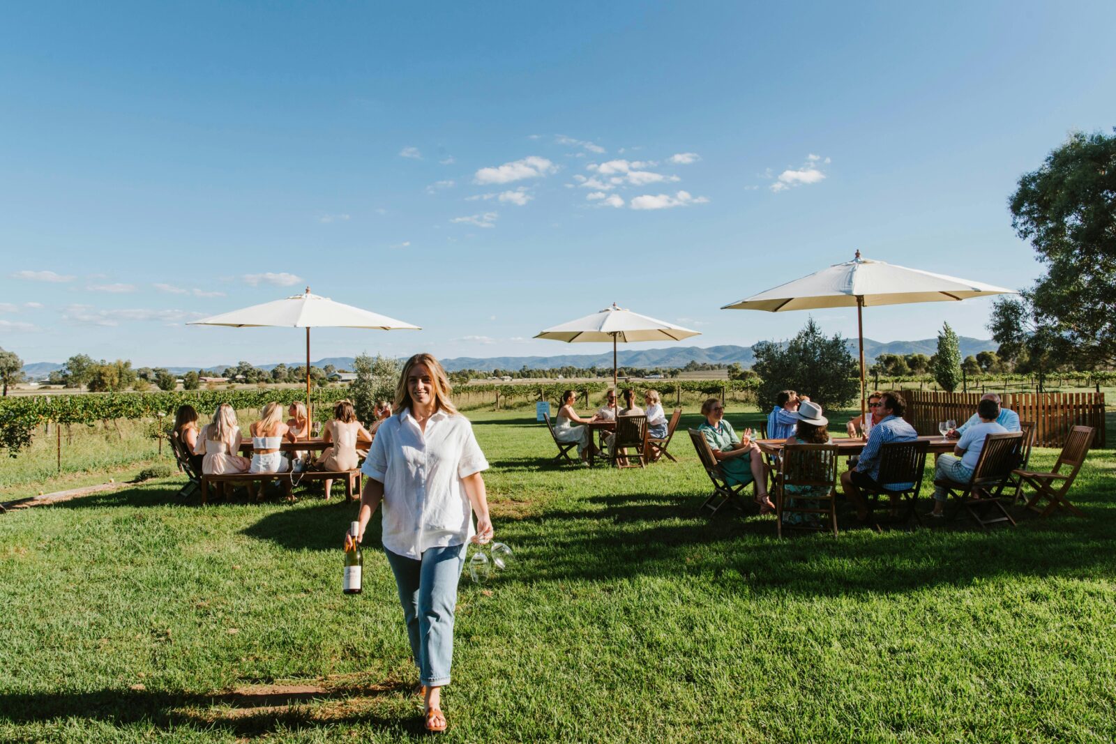 Woman walking toward the camera holding a bottle of wine and glasses. Tables of people in background