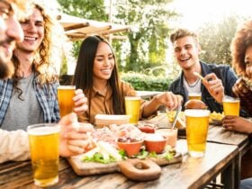 young people around a table in the outdoors smiling and enjoying beers and a platter of food