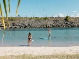 Man stand up paddleboarding and lady walking into blue water