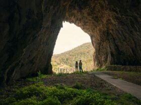A couple seen from a distance at the entrance of a cave, with a bright sunny landscape ahead of them