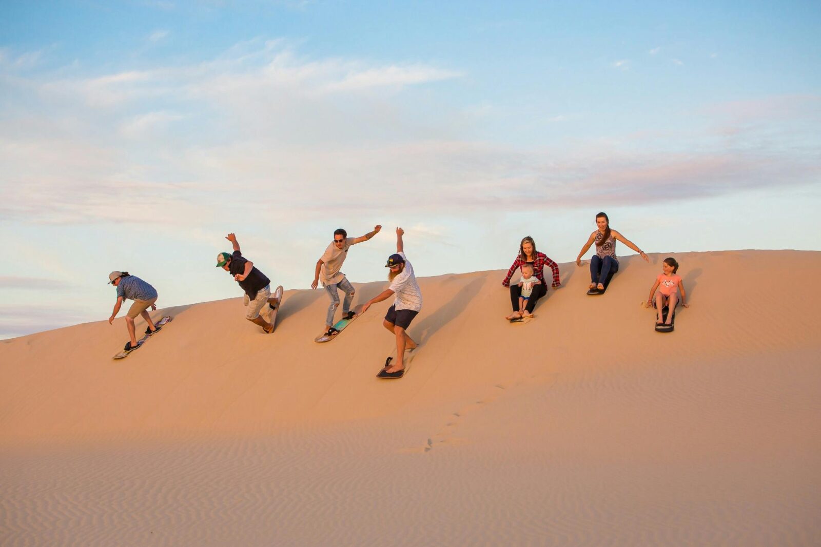 group of people of all different ages sandboarding down the sand dunes