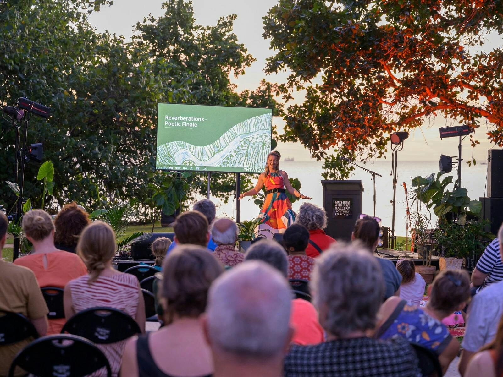 A woman on stage, screen saying NT Writers Festival. Audience is looking at ocean backdrop