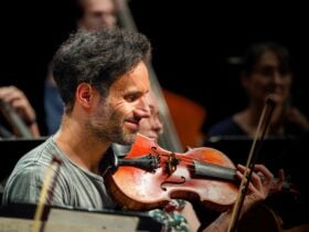 A man sits in the orchestra with his viola while smiling