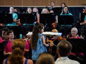 A young girl stands in front of an orchestra with a conductors baton, smiling.