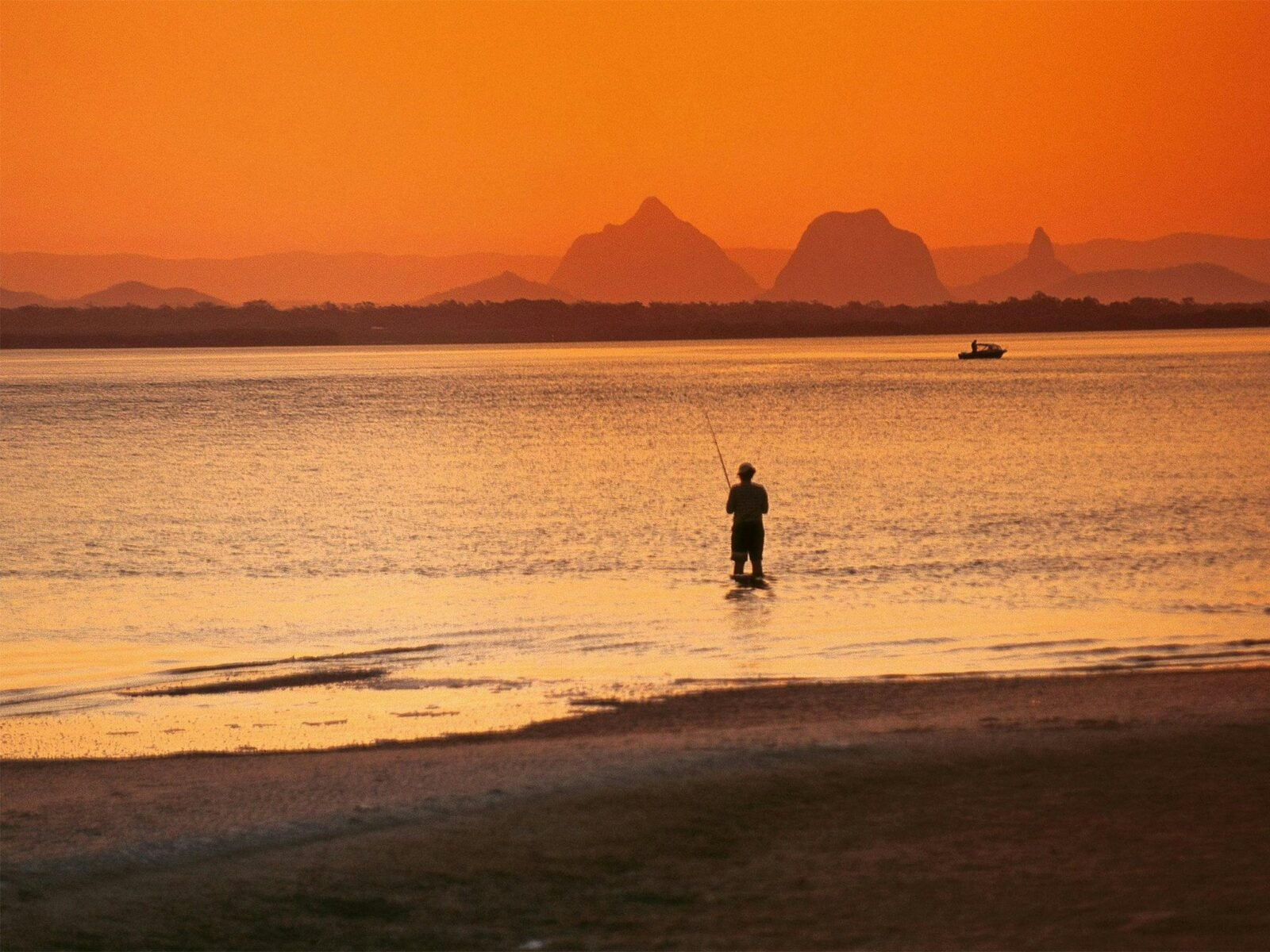 Oranged hued image of person standing in the ocean fishing with a boat further out and mountains