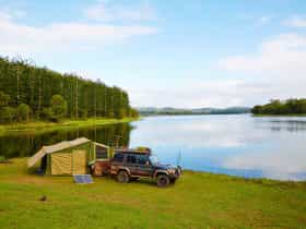 Tent and car are set on the lakes shores with pine forest in background.