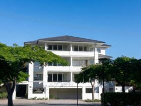 A white three-storey apartment building with balconies on each level, framed by mature trees, with a