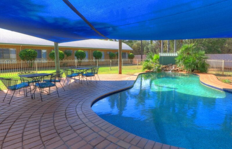 Photo shows pool and sitting area with waterfall, trees and lawns and motel rooms in background