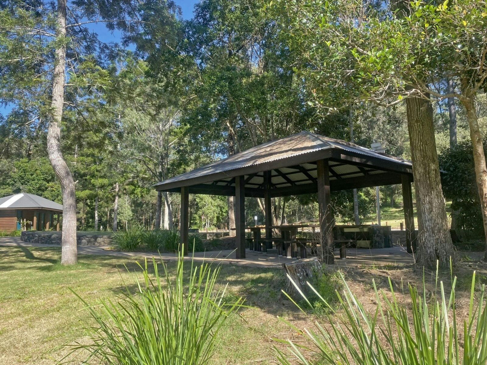 Sheltered picnic tables in grassy area with a toilet block in the background.