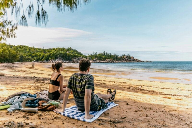 Two people sitting on towels on the beach looking out at a bay.