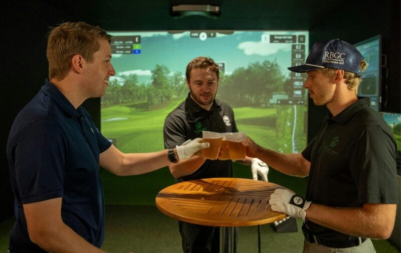 3 men enjoy beers and cheers to a great round in front of the golf simulator