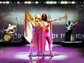 Two women standing back to back in 70s era clothing - white boots and gold capes