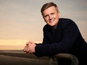 Aled Jones stands facing the camera with his hands clasped together smiling wearing a black jumper