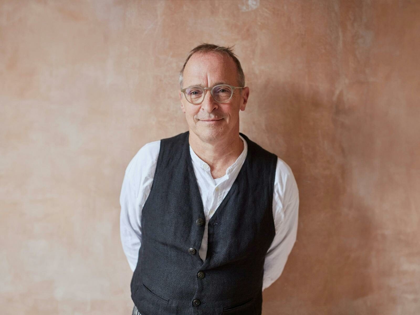 Man smiles at camera wearing white long sleeve with dark vest and glasses