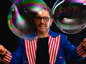 Man with short dark hair and glasses, red and white striped vest on with bubbles in front of him