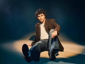 A young man with dark curly hair sitting on the floor in brown jacket and jeans
