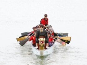 A dragon boat crew paddling toward the camera on calm water, led by a drummer at the front.
