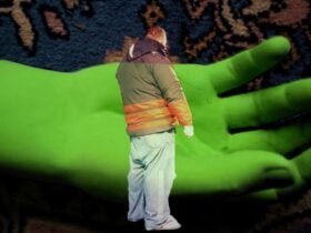 Video still of a man in a jacket standing in front of a large green hand