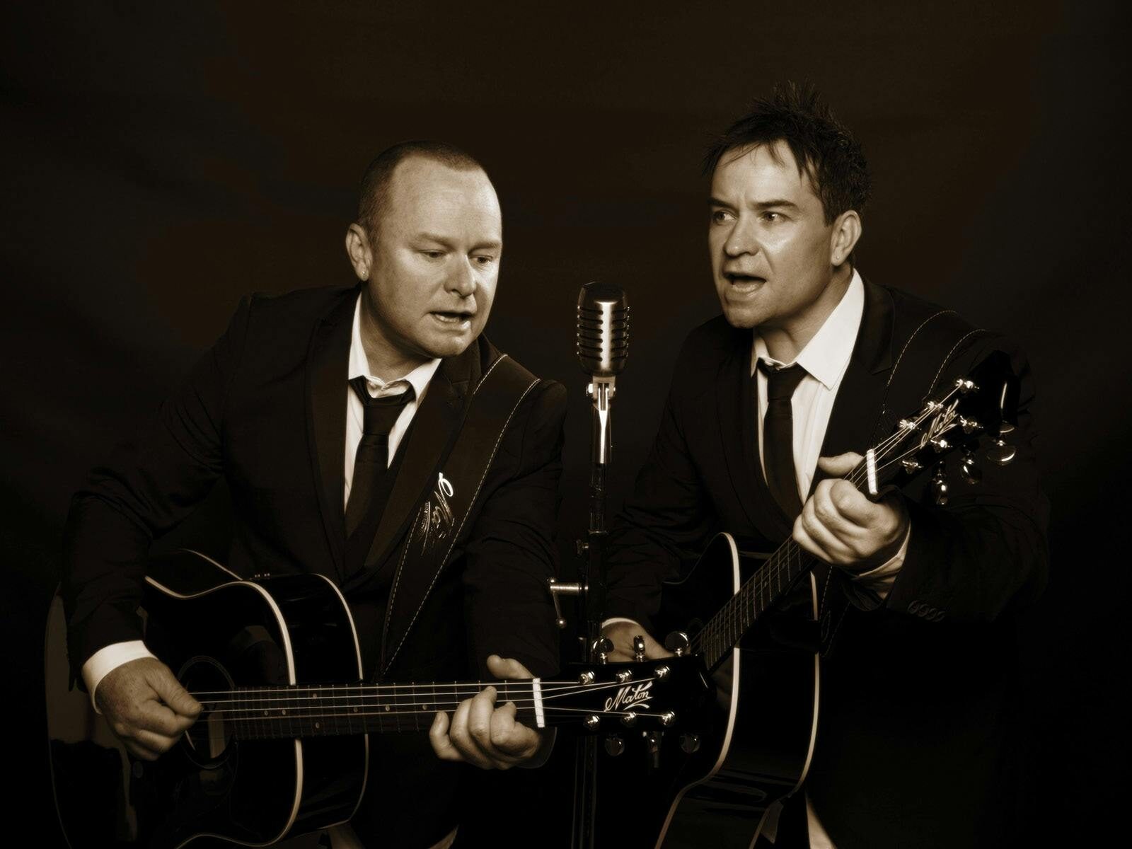 2 men dressed in dark suits, white shirts and ties playing guitars and singing into a microphone
